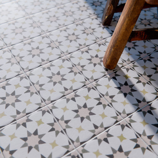 Marketing image of Agadir Natural Moroccan-style 20x20cm porcelain tiles laid on a floor, featuring a soft neutral geometric star pattern with subtle grey and warm beige tones, styled with a wooden chair for a lived-in look.