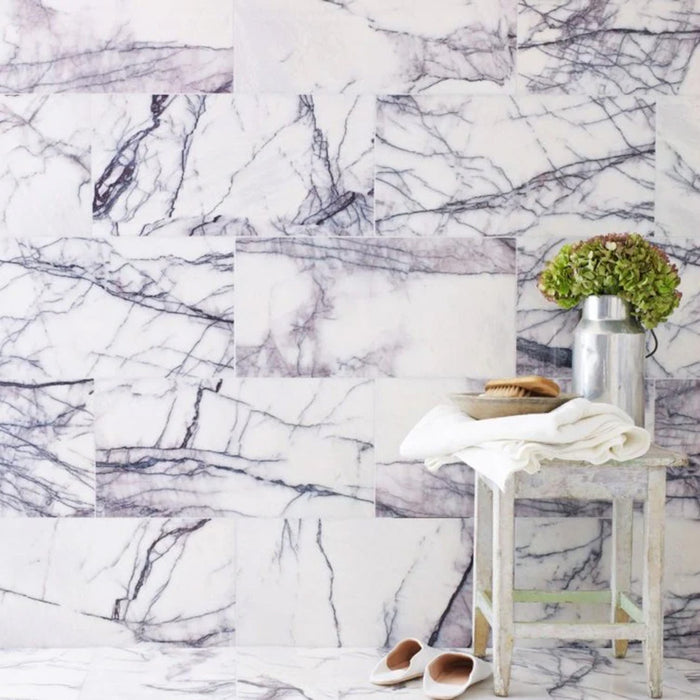 Bathroom interior featuring Fayrouz Lilac 30.5x61cm natural marble tiles with a honed finish, installed across walls and floor in a staggered layout, showcasing a soft white base with flowing lilac and grey veining, styled with a rustic wooden stool, brushed metal vase with greenery, and neutral accessories for a calm, refined space.