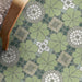 Nantes Green Moroccan-style tiles in a 20x20cm format, laid as a decorative patterned floor. The repeating floral motif in soft green tones is complemented by mosaic-style detailing in muted neutrals and white, styled with a wooden chair to showcase the tiles in a warm, characterful interior setting.