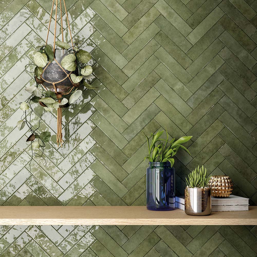 Nomad Sage Green brick wall tiles in a 6 × 24.6cm format, laid in a herringbone pattern with a glossy, lightly textured finish, creating a rich green industrial-style feature wall styled with plants and shelving.