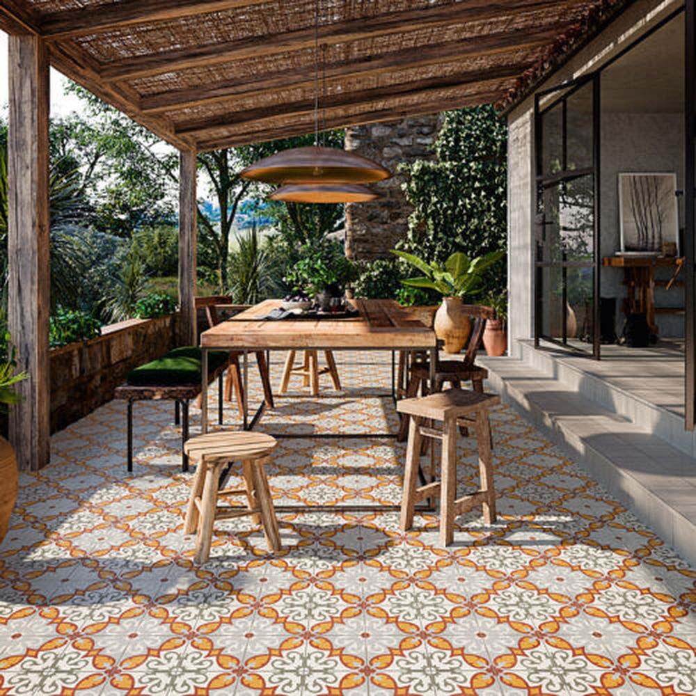 Rustic outdoor dining area featuring Mediterraneo Gela 2cm patterned outdoor slabs 50 × 100cm, showcasing a decorative Mediterranean-inspired pattern in warm terracotta, grey, and white tones, paired with natural wood furniture and lush greenery.