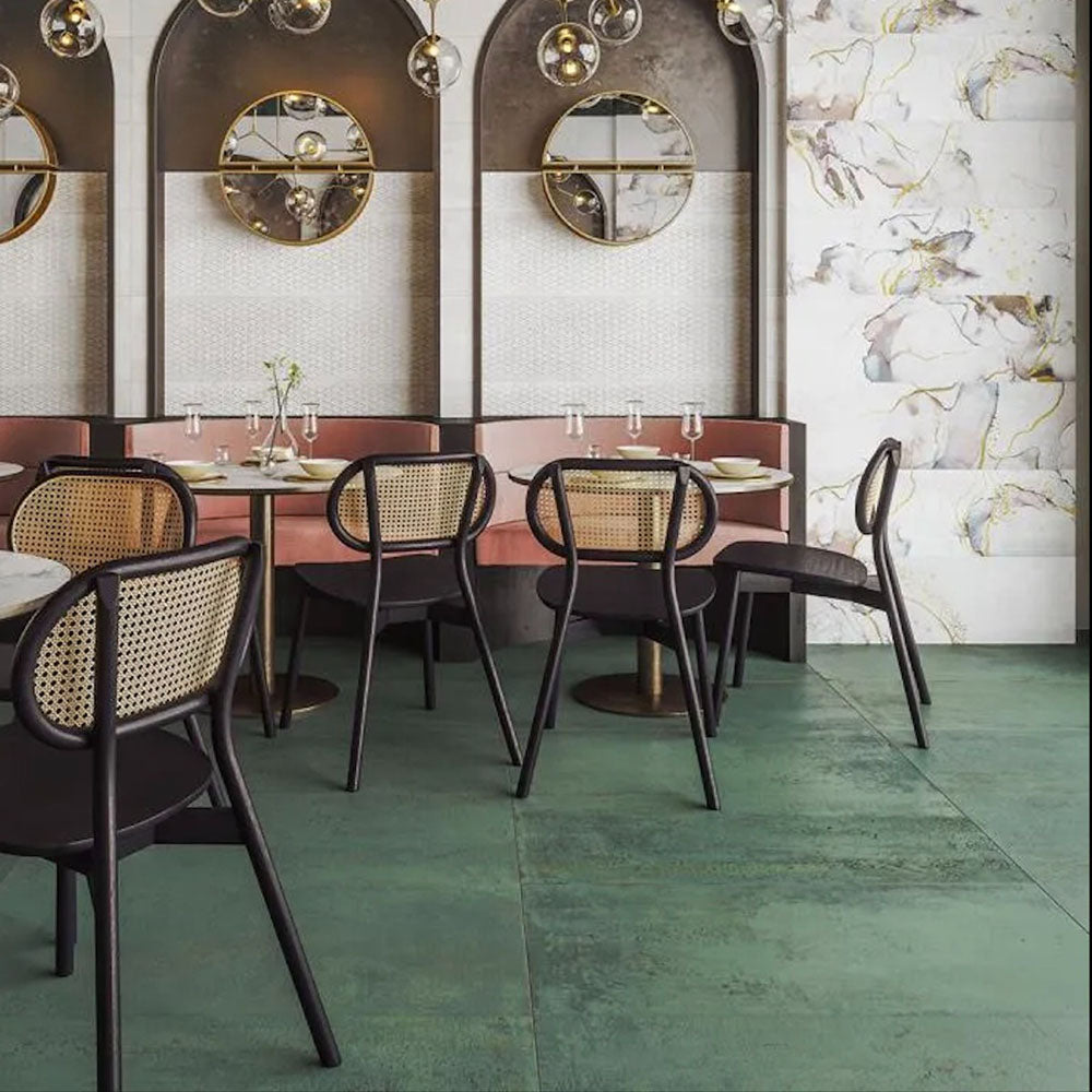 Contemporary restaurant interior featuring Mediterraneo Green 2cm outdoor slabs 50 × 100cm, showcasing deep green stone-effect porcelain tiles, paired with cane dining chairs, brass lighting, and floral marble-effect feature walls.