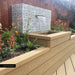 Garden water feature featuring Mediterraneo Olbia 2cm Patterned Outdoor Slabs 50x100cm, showcasing a decorative Mediterranean-style pattern in soft green, blue and terracotta tones, surrounded by timber decking, raised planters and colourful planting.