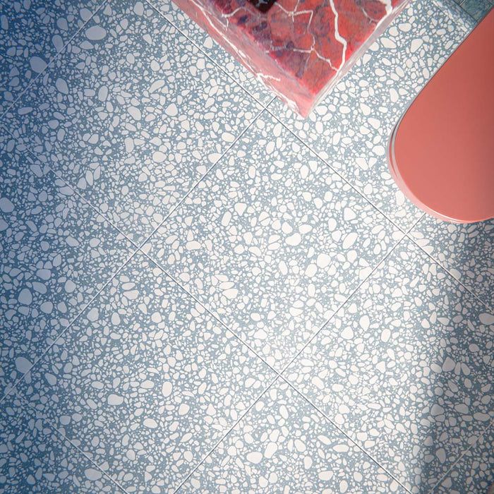 Eclectic bathroom featuring Melody POP Terrazzo Blue 60 × 60cm terrazzo-effect porcelain floor tiles, showcasing soft white stone fragments scattered across a muted blue base, paired with glossy blue wall tiles, a dramatic red marble-effect basin, and playful contemporary styling.