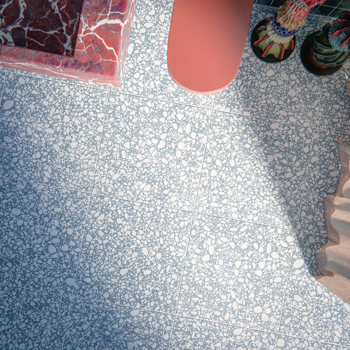 Eclectic bathroom featuring Melody POP Terrazzo Blue 60 × 60cm terrazzo-effect porcelain floor tiles, showcasing soft white stone fragments scattered across a muted blue base, paired with glossy blue wall tiles, a dramatic red marble-effect basin, and playful contemporary styling.