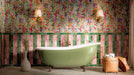 Bathroom featuring Melody POP Terrazzo Green 60 × 60cm terrazzo-effect porcelain floor tiles, showcasing a playful green and pink speckled design, paired with striped glossy wall tiles, floral wallpaper, and a sage green freestanding bath for a bold maximalist-inspired interior.