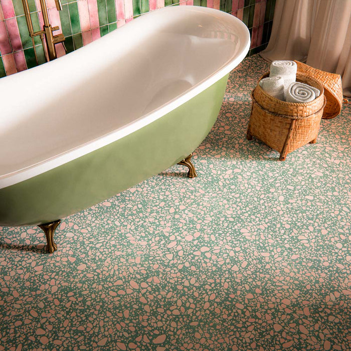 Bathroom featuring Melody POP Terrazzo Green 60x60cm terrazzo effect floor tiles, with a green background and pink speckled detailing, paired with pink and green wall tiles, colourful floral wallpaper and a freestanding green bathtub with brass accents.