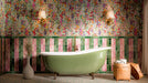 Bathroom featuring Melody POP Terrazzo Green 60x60cm terrazzo effect floor tiles, with a green background and pink speckled detailing, paired with pink and green wall tiles, colourful floral wallpaper and a freestanding green bathtub with brass accents.