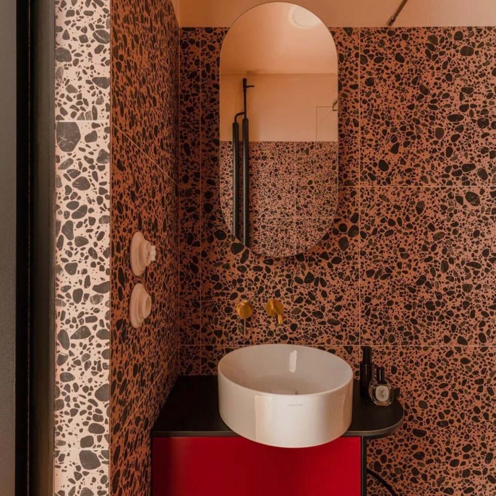 Bold bathroom interior featuring Melody POP Terrazzo Pink 60 × 60cm porcelain tiles, showcasing a striking terrazzo-effect design with oversized dark stone fragments set against warm pink tones, paired with black fittings and vibrant red vanity units for a playful contemporary look.