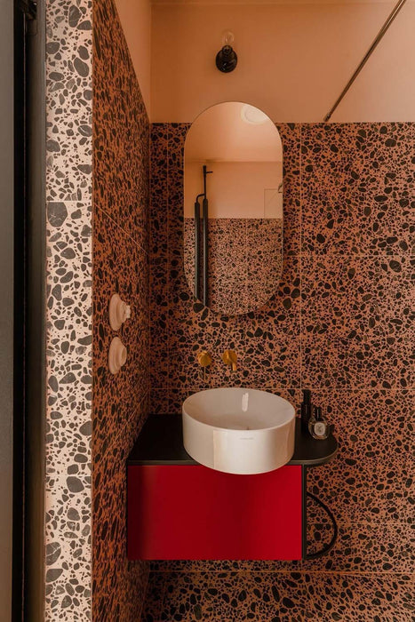 Bold bathroom interior featuring Melody POP Terrazzo Pink 60 × 60cm porcelain tiles, showcasing a striking terrazzo-effect design with oversized dark stone fragments set against warm pink tones, paired with black fittings and vibrant red vanity units for a playful contemporary look.