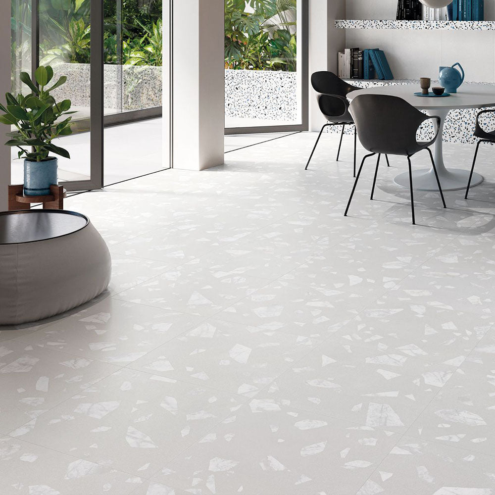 Open-plan interior featuring Melody ROCK Terrazzo White 60 × 60cm terrazzo-effect porcelain floor tiles, showcasing oversized marble-inspired fragments on a crisp white base, paired with minimalist black furniture, soft neutral tones, and large glazing for a bright contemporary setting.