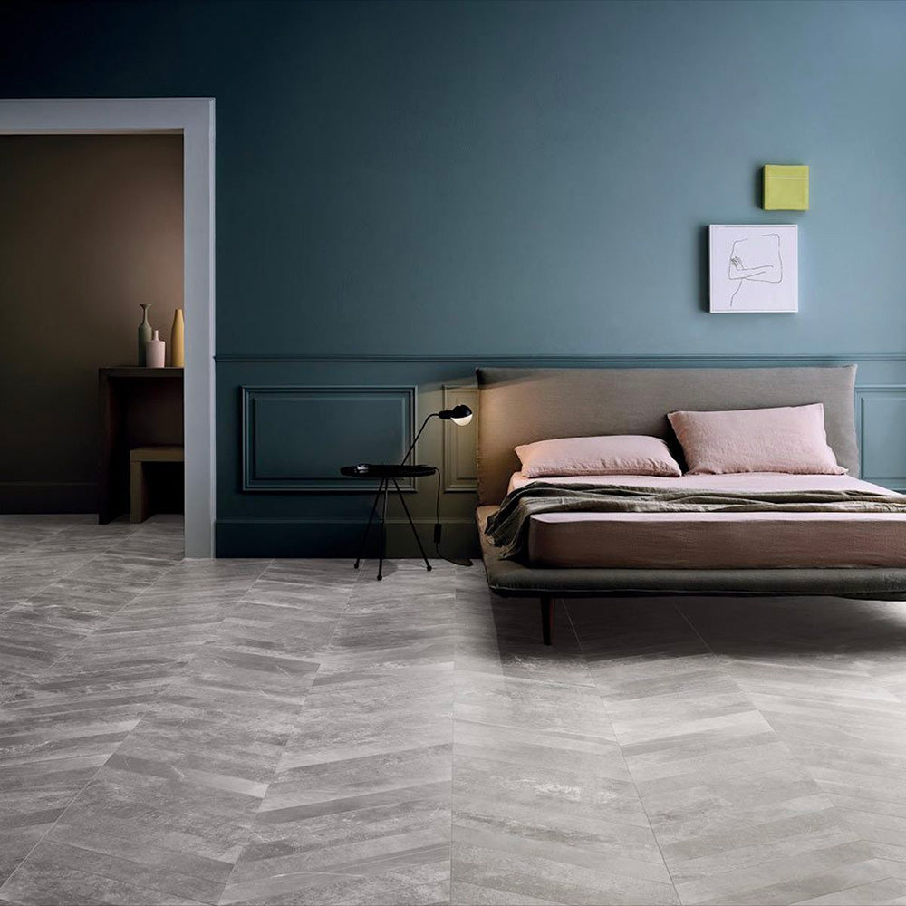 Bedroom featuring Monzo Stone Chevron Titanium 10x53cm stone-effect porcelain chevron floor tiles in soft grey tones, styled with deep blue walls, modern furniture, and subtle natural-style veining for a contemporary interior look.