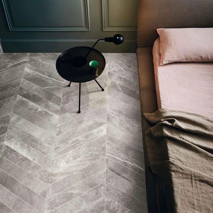 Bedroom featuring Monzo Stone Chevron Titanium 10x53cm stone-effect porcelain chevron floor tiles in soft grey tones, styled with deep blue walls, modern furniture, and subtle natural-style veining for a contemporary interior look.