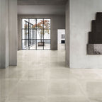 Lifestyle image of Monzo Stone Sand 60x60cm polished stone-effect porcelain floor tiles in soft warm beige tones, featuring delicate veining and subtle texture, styled within a spacious modern interior with minimalist architecture and large black-framed windows.