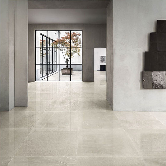 Lifestyle image of Monzo Stone Sand 60x60cm polished stone-effect porcelain floor tiles in soft warm beige tones, featuring delicate veining and subtle texture, styled within a spacious modern interior with minimalist architecture and large black-framed windows.