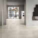 Lifestyle image of Monzo Stone Sand 60x60cm polished stone-effect porcelain floor tiles in soft warm beige tones, featuring delicate veining and subtle texture, styled within a spacious modern interior with minimalist architecture and large black-framed windows.