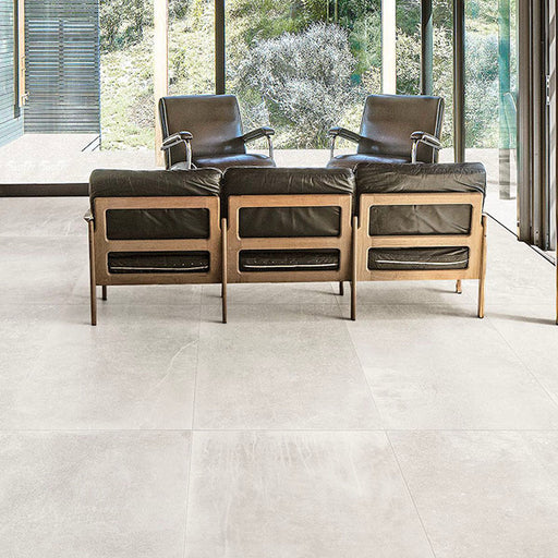 Living room featuring Monzo Stone Silk 60x60cm stone-effect porcelain floor tiles in soft ivory tones, styled with black leather lounge chairs, warm wood furniture, and floor-to-ceiling glazing for a modern interior look.