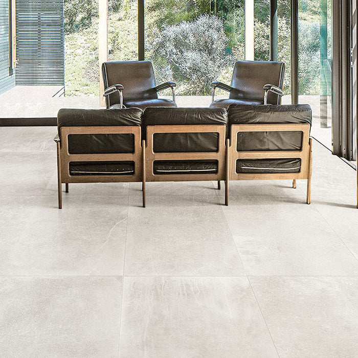 Living room featuring Monzo Stone Silk 60x60cm stone-effect porcelain floor tiles in soft ivory tones, styled with black leather lounge chairs, warm wood furniture, and floor-to-ceiling glazing for a modern interior look.