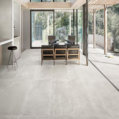 Living room featuring Monzo Stone Silver 60x60cm stone-effect porcelain floor tiles in soft silver-grey tones, styled with black leather seating, natural wood finishes, and floor-to-ceiling glazing for a contemporary interior look.