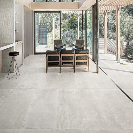 Living room featuring Monzo Stone Silver 60x60cm stone-effect porcelain floor tiles in soft silver-grey tones, styled with black leather seating, natural wood finishes, and floor-to-ceiling glazing for a contemporary interior look.