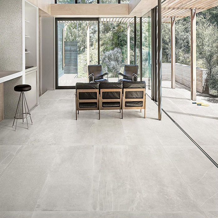 Living room featuring Monzo Stone Silver 60x60cm stone-effect porcelain floor tiles in soft silver-grey tones, styled with black leather seating, natural wood finishes, and floor-to-ceiling glazing for a contemporary interior look.