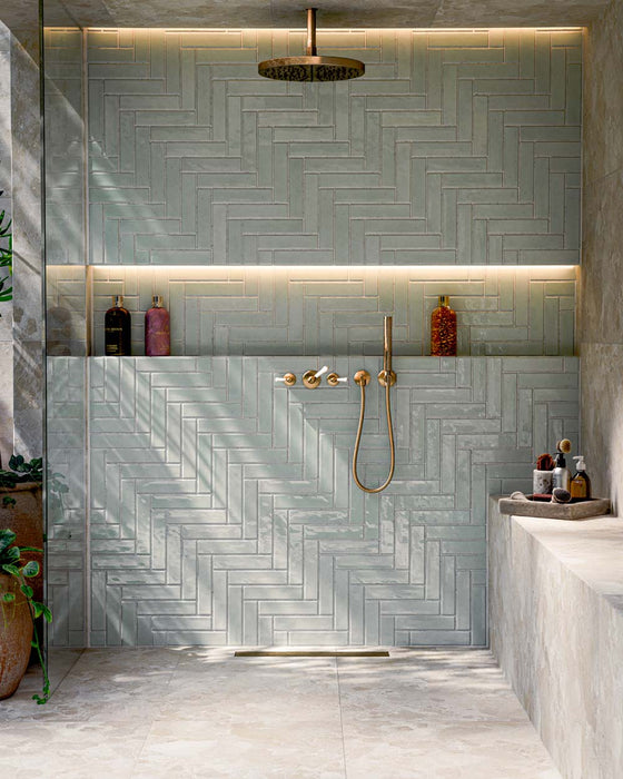 Bathroom featuring Nomad Mint Green Brick 6x24.6cm industrial-style wall tiles, laid in a herringbone pattern and showcasing a glossy mint green finish, paired with brass shower fittings, recessed shelving and warm ambient lighting.