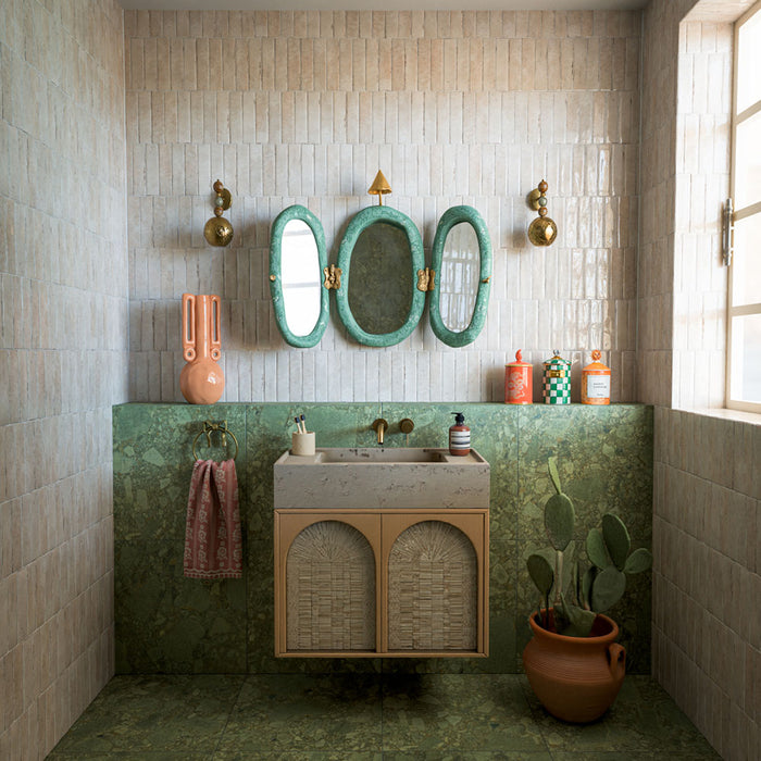 Bathroom featuring Nomad Oatmeal industrial style wall tiles, showcasing soft oatmeal and beige tones with subtle surface variation and a glossy finish. Installed in a vertical stacked layout, the tiles create texture and depth while complementing the rich green stone-effect surfaces, natural materials and brass accents. The result is a warm, characterful space that blends contemporary design with earthy Mediterranean-inspired styling.
