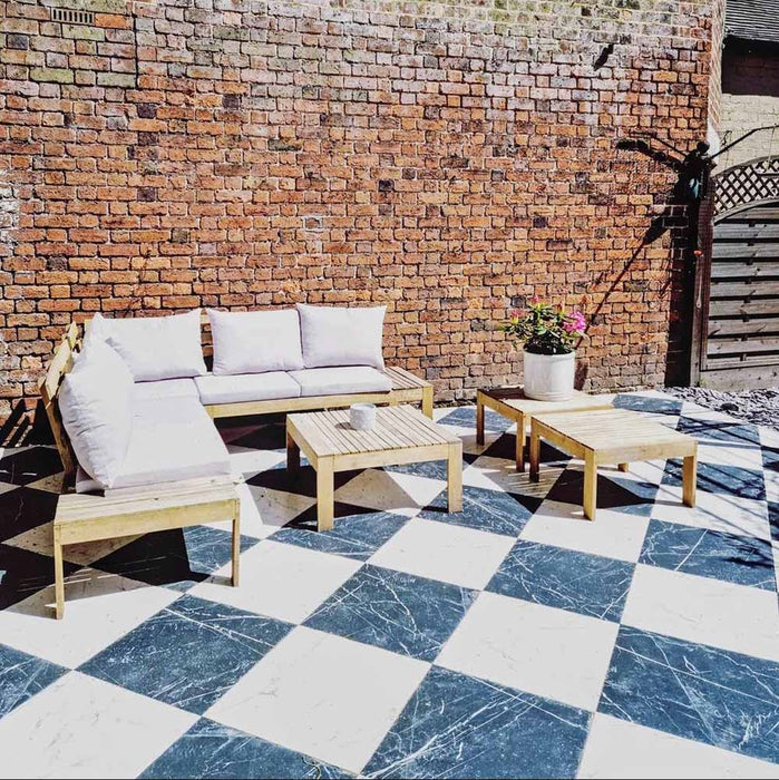 Outdoor seating area featuring Florence Grand Calacatta White 50x50cm marble-effect porcelain floor tiles laid in a classic black and white checkerboard pattern, styled with wooden garden furniture, soft cushions and exposed brick walls for a relaxed yet elegant outdoor space.