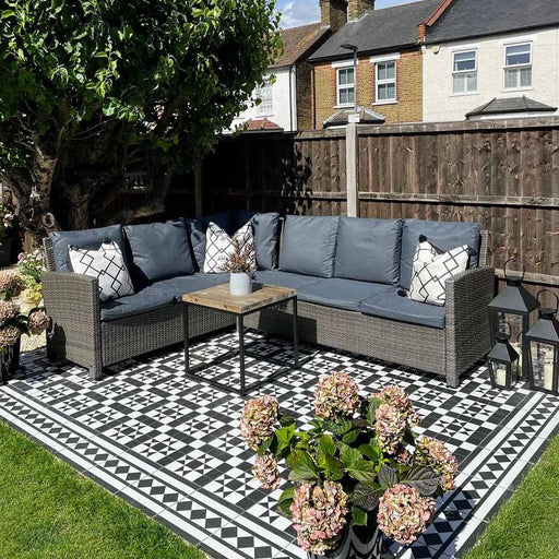 Outdoor seating area featuring Dorset Tile Black & White Victorian-style porcelain floor tiles in a 25 × 25cm format, laid in a bold geometric pattern with a decorative border, styled with woven garden furniture, lantern lighting and planting to create a classic yet contemporary garden space.