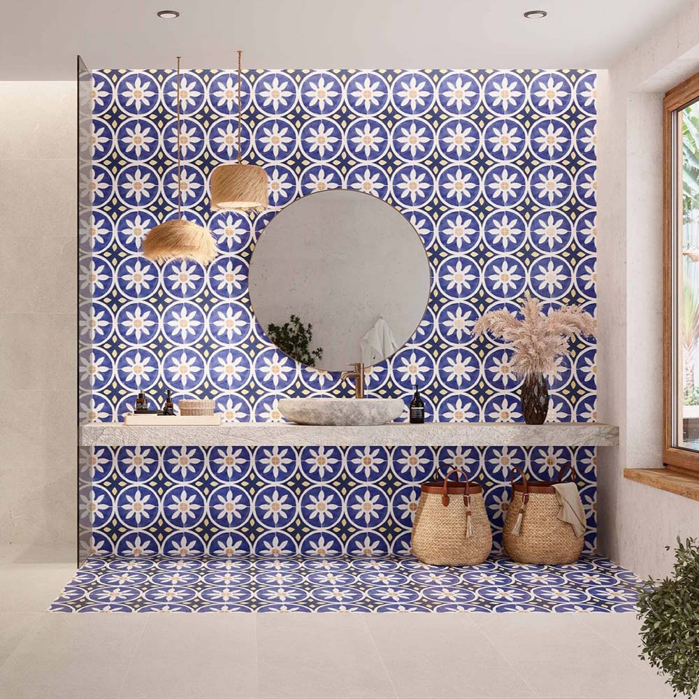 Bathroom featuring Peniscola Blue and Yellow patterned wall and floor tiles, showcasing a bold Mediterranean-inspired floral geometric design in vibrant cobalt blue, warm yellow and crisp white tones, paired with a floating stone vanity, round mirror, woven pendant lights, natural baskets and soft neutral finishes for a bright, characterful interior.