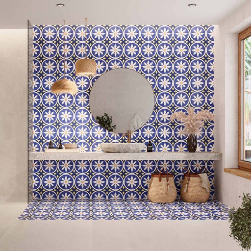 Bathroom featuring Peniscola Blue and Yellow patterned wall and floor tiles, showcasing a bold Mediterranean-inspired floral geometric design in vibrant cobalt blue, warm yellow and crisp white tones, paired with a floating stone vanity, round mirror, woven pendant lights, natural baskets and soft neutral finishes for a bright, characterful interior.