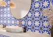 Bathroom featuring Peniscola Blue and Yellow patterned wall and floor tiles, showcasing a bold Mediterranean-inspired floral geometric design in vibrant cobalt blue, warm yellow and crisp white tones, paired with a floating stone vanity, round mirror, woven pendant lights, natural baskets and soft neutral finishes for a bright, characterful interior.