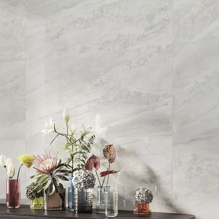 Living room featuring Petra Avorio Ivory stone marble effect porcelain wall tiles in a 60cm x 120cm format, showcasing soft ivory, pearl white and light grey veining inspired by natural stone, paired with colourful glass vases, fresh floral arrangements and contemporary styling to create a bright, elegant interior.