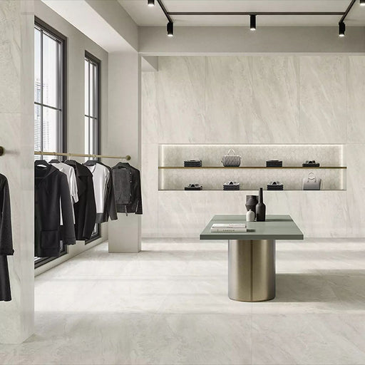 Retail showroom featuring Petra Beige stone marble effect porcelain wall and floor tiles in a 30cm x 60cm format, showcasing warm beige, ivory and soft cream veining inspired by natural stone, paired with minimalist shelving, contemporary display furniture, black metal clothing rails and a bright, modern commercial interior.