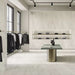Retail showroom featuring Petra Beige stone marble effect porcelain wall and floor tiles in a 30cm x 60cm format, showcasing warm beige, ivory and soft cream veining inspired by natural stone, paired with minimalist shelving, contemporary display furniture, black metal clothing rails and a bright, modern commercial interior.