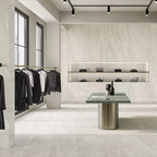 Retail showroom featuring Petra Beige stone marble effect porcelain wall and floor tiles in a 60cm x 120cm format, showcasing warm beige, ivory and soft cream veining inspired by natural stone, paired with minimalist shelving, contemporary display furniture, black metal clothing rails and a bright, modern commercial interior.