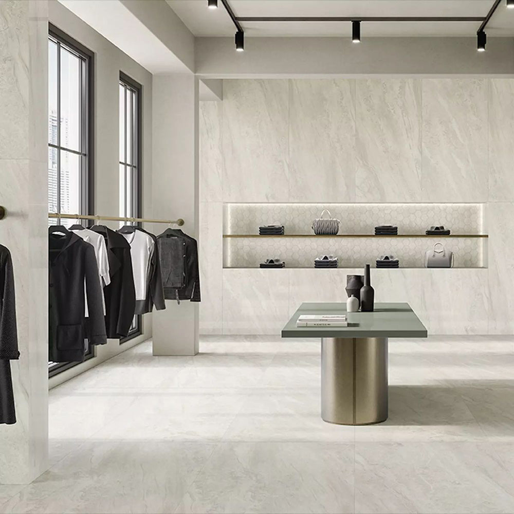 Retail showroom featuring Petra Beige stone marble effect porcelain wall and floor tiles in a 60cm x 120cm format, showcasing warm beige, ivory and soft cream veining inspired by natural stone, paired with minimalist shelving, contemporary display furniture, black metal clothing rails and a bright, modern commercial interior.