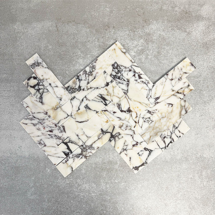 Arrangement featuring Fayrouz Picasso 5x20cm natural marble tiles with a honed finish, laid in a herringbone pattern to create directional movement, showcasing a white base with bold grey, green and warm gold veining and natural variation across each piece.