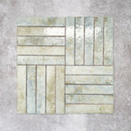 Product display of Pottery Brick Agave Green 5x25cm glossy wall tiles in soft green and sage tones, arranged in a basketweave layout with handcrafted edges and subtle tonal variation against a textured concrete-style backdrop.