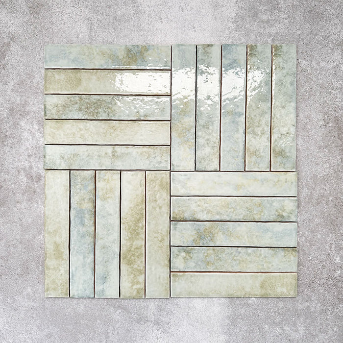 Product display of Pottery Brick Agave Green 5x25cm glossy wall tiles in soft green and sage tones, arranged in a basketweave layout with handcrafted edges and subtle tonal variation against a textured concrete-style backdrop.