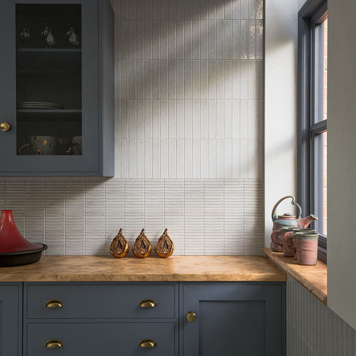 Kitchen splashback featuring Pottery Decor Blanco 12.5 × 25cm ceramic wall tiles, combining textured linear relief tiles with glossy slim brick tiles in soft white tones, paired with deep blue cabinetry, warm wood worktops, and brass accents for a layered contemporary design.