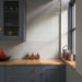 Kitchen splashback featuring Pottery Decor Blanco 12.5 × 25cm ceramic wall tiles, combining textured linear relief tiles with glossy slim brick tiles in soft white tones, paired with deep blue cabinetry, warm wood worktops, and brass accents for a layered contemporary design.