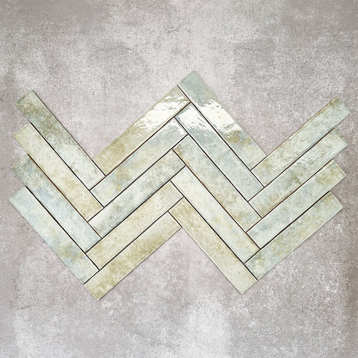 Product display of Pottery Brick Agave Green 5x25cm glossy wall tiles in soft green and sage tones, arranged in a herringbone layout with handcrafted edges and subtle tonal variation against a textured concrete-style backdrop.