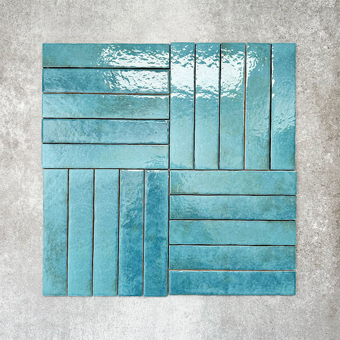 Product display of Pottery Brick Azul Blue 5x25cm glossy wall tiles in vibrant blue tones, arranged in a basketweave layout with handcrafted edges and tonal variation against a textured concrete-style backdrop.