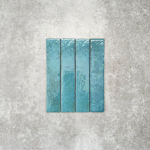 Product display of Pottery Brick Azul Blue 5x25cm glossy wall tiles in vibrant blue tones, featuring handcrafted edges and subtle tonal variation against a textured concrete-style backdrop.