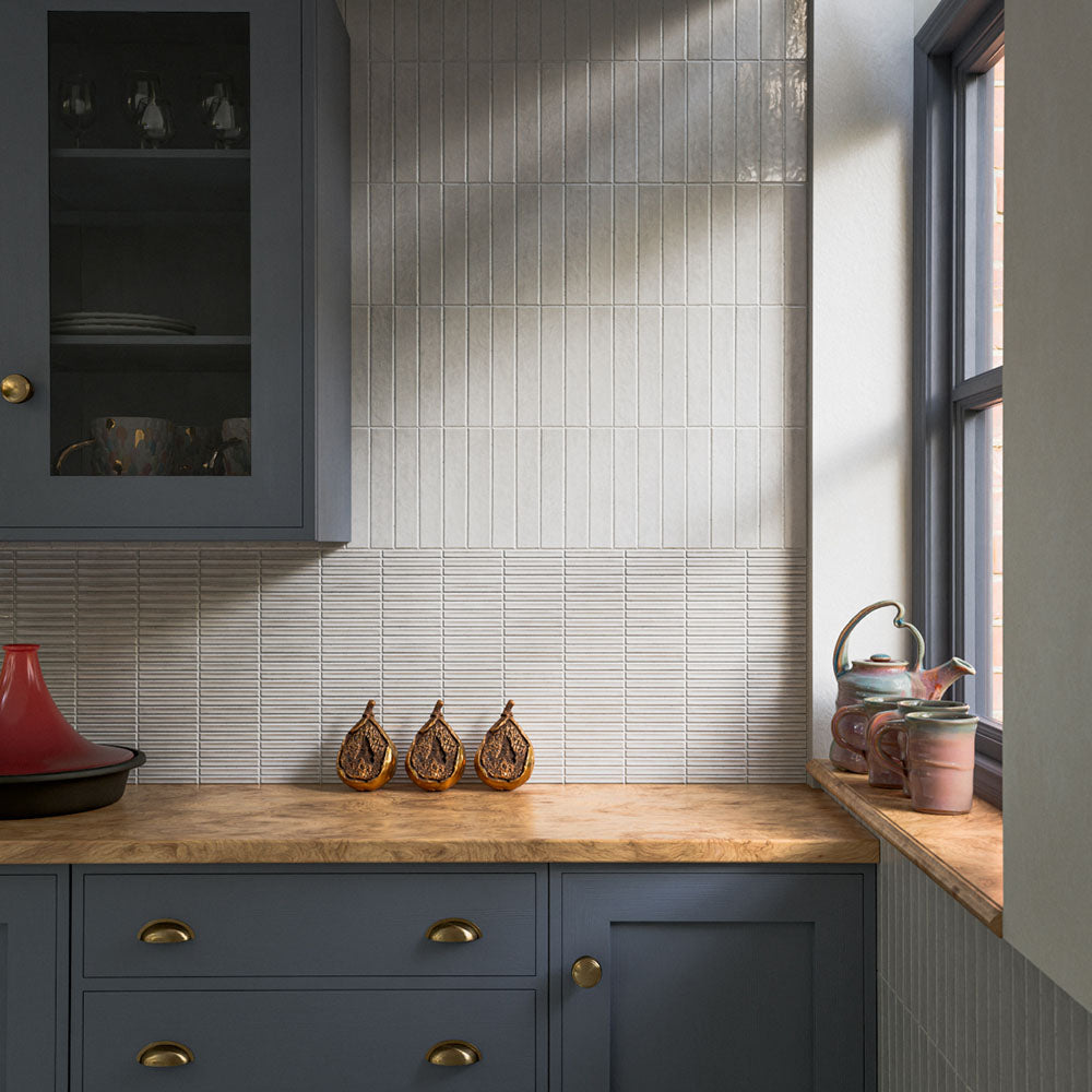 Kitchen splashback featuring Pottery Brick Blanco 5 × 25cm ceramic wall tiles, showcasing soft tonal variation, a glossy handcrafted finish, and a slim brick format, paired with deep blue cabinetry, warm wood worktops, and brass accents for a refined contemporary look.