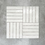Product display of Pottery Brick Blanco 5x25cm glossy wall tiles in soft white tones, arranged in a basketweave layout with handcrafted edges and subtle surface variation against a textured concrete-style backdrop.