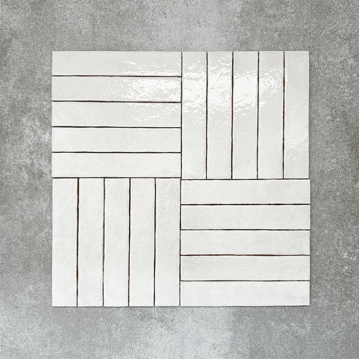 Product display of Pottery Brick Blanco 5x25cm glossy wall tiles in soft white tones, arranged in a basketweave layout with handcrafted edges and subtle surface variation against a textured concrete-style backdrop.
