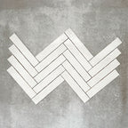 Product display of Pottery Brick Blanco 5x25cm glossy wall tiles in soft white tones, arranged in a herringbone layout with handcrafted edges and subtle surface variation against a textured concrete-style backdrop.