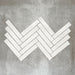 Product display of Pottery Brick Blanco 5x25cm glossy wall tiles in soft white tones, arranged in a herringbone layout with handcrafted edges and subtle surface variation against a textured concrete-style backdrop.